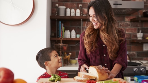 A mother smiles as she slices a loaf of bread on a counter and looks down at her son who is smiling back at her.
