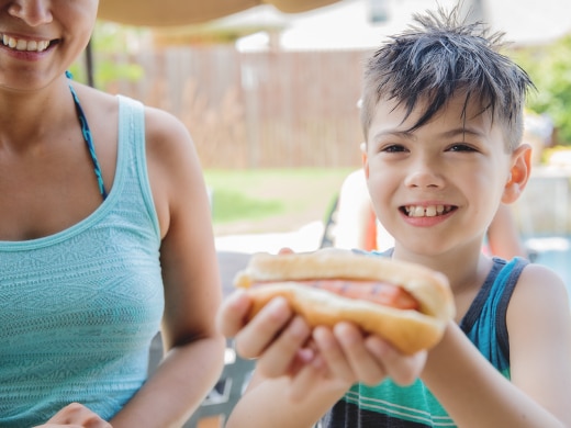 A Mom and a boy at a pool. The smiling boy is being handed a hot dog.